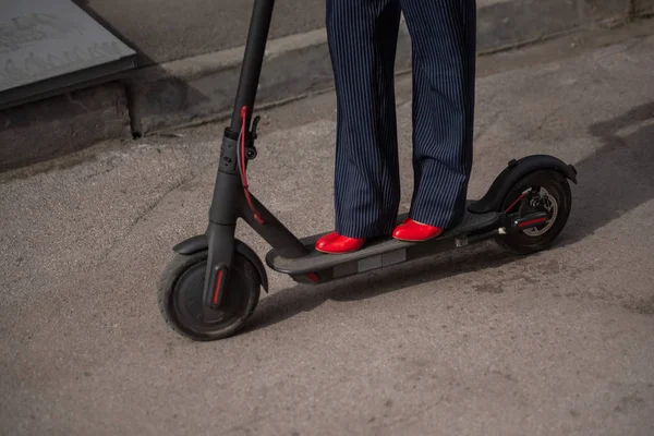 Młoda kobieta w odzieży formalnej na czerwonym obcasie stoi na skuter elektryczny. Zbliżenie kobiecych nóg. Kobieta biznesowa w garniturze spodni i czerwone buty porusza się po mieście na skuterze elektrycznym. — Zdjęcie stockowe