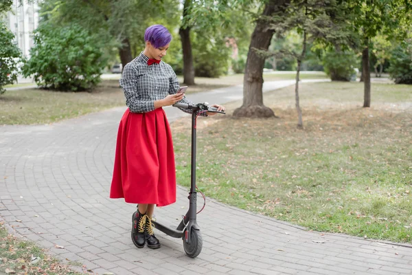 Młoda kobieta z fioletowymi włosami jeździ skuter elektryczny w parku. Stylowa dziewczyna z ogoloną świątynią w szachownicę, długa czerwona spódnica i muszka pisze wiadomość na smartfonie. Hipster — Zdjęcie stockowe