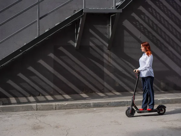 Rudowłosy dziewczyna w białej koszulce napędza skuter elektryczny wzdłuż ściany. Kobieta biznesu w garniturze spodni i czerwone wysokie obcasy jeździ po mieście w nowoczesnym samochodzie. Kobieta biznesu na skuterze. — Zdjęcie stockowe