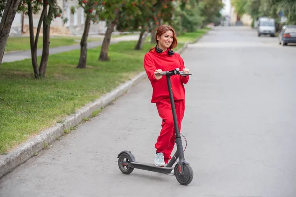 Рыжая девушка в красном спортивном костюме водит электрический скутер. Молодая женщина в огромной одежде ездит по городу на современном транспорте и слушает музыку с помощью беспроводных наушников . — стоковое фото