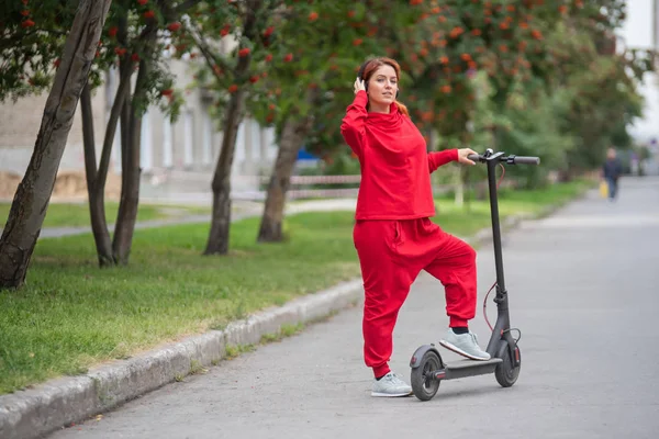 Menina de cabelos vermelhos em um traje vermelho dirige uma scooter elétrica. Uma jovem mulher de roupas grandes passeia pela cidade em transportes modernos e ouve música usando fones de ouvido sem fio . — Fotografia de Stock