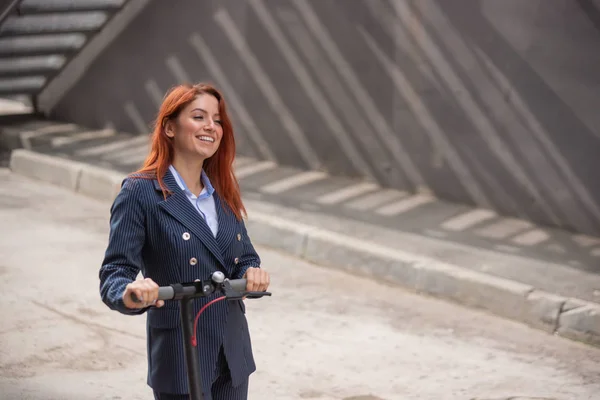 A red-haired girl in a jacket and trousers drives an electric scooter along the wall. A business woman in a pantsuit and red high heels rides around the city in a modern car. Office worker. Dress code