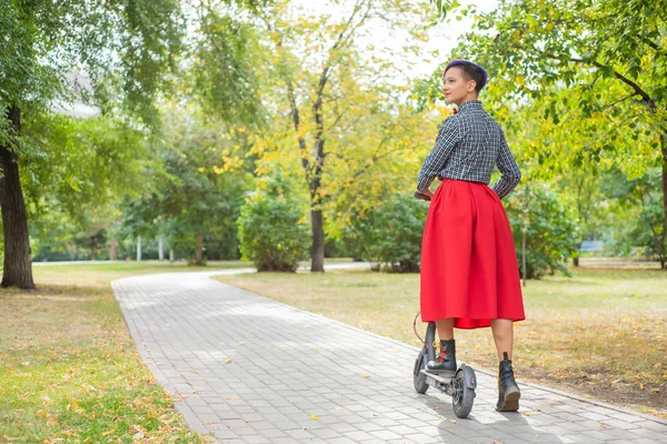 Une jeune femme aux cheveux violets conduit un scooter électrique dans un parc. Une fille élégante avec un temple rasé dans une chemise à carreaux, une longue jupe rouge et un nœud papillon se promène dans la ville sur un appareil moderne . — Photo