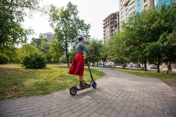 A young woman with purple hair rides an electric scooter in a park. A stylish girl with a shaved temple in a plaid shirt, a long red skirt and a bow tie is riding around the city on a modern device.