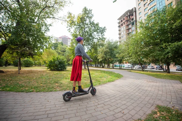 Egy fiatal nő, lila hajú lovagol egy elektromos robogó egy parkban. Egy stílusos lány egy borotvált templom egy kockás ing, egy hosszú vörös szoknya és egy csokornyakkendő a lovaglás a város körül egy modern eszköz. — Stock Fotó