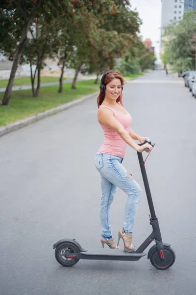 Une jeune femme rousse en jeans déchiré et sandales à talons hauts monte un scooter électrique sur la route et écoute de la musique sur son casque sans fil bluetooth. Transport électrique pratique . — Photo