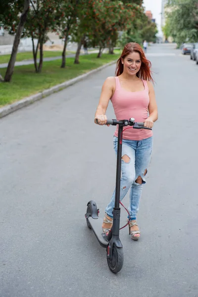 Una joven pelirroja en pantalones vaqueros rasgados y sandalias de tacón alto monta una scooter eléctrica en la carretera. Vehículos eléctricos convenientes para un movimiento rápido por la ciudad . —  Fotos de Stock