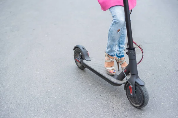 Gros plan des jambes des femmes en jeans déchiré et sandales à talons hauts à côté d'un scooter électrique. Une fille dans une veste rose se déplace rapidement dans la ville sur un moyen de transport électrique pratique . — Photo