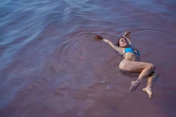 En kvinna i en blå bikini simmar på ryggen i en Saltsjö. Mirakel av naturen rosa sjön. Deponering och extraktion av salt. Tätt vatten som skjuter till ytan. Terapeutiska ingrepp i mineralvatten. — Stockfoto