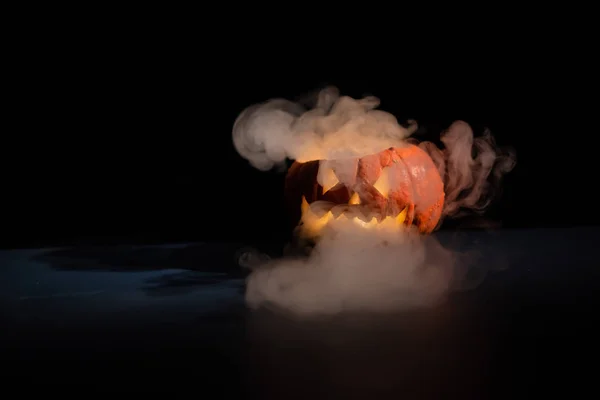 Halloween, oranje pompoen met een eng licht gezicht op een donkere achtergrond. Dikke grijze rook komt naar buiten en verspreidt zich over de zwarte tafel. Een close-up van een zaklamp aan de vooravond van alle heiligen — Stockfoto