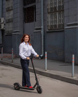 Beyaz gömlekli kızıl saçlı bir kız duvar boyunca elektrikli scooter kullanıyor. Pantolon kostümlü ve kırmızı topuklu ayakkabıgiyen bir iş kadını modern bir arabayla şehirde dolaşıyor. ofiste kıyafet kodu.