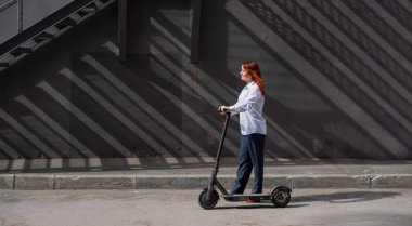 Beyaz gömlekli kızıl saçlı bir kız duvar boyunca elektrikli scooter kullanıyor. Pantolon kostümlü ve kırmızı topuklu ayakkabıgiyen bir iş kadını modern bir arabayla şehirde dolaşıyor. Scooter'da iş kadını.