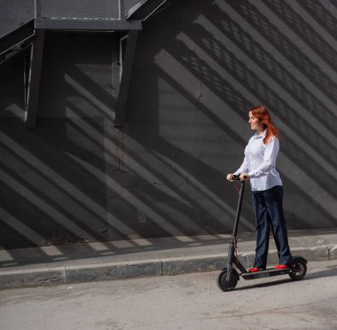Beyaz gömlekli kızıl saçlı bir kız duvar boyunca elektrikli scooter kullanıyor. Pantolon kostümlü ve kırmızı topuklu ayakkabıgiyen bir iş kadını modern bir arabayla şehirde dolaşıyor. Scooter'da iş kadını.