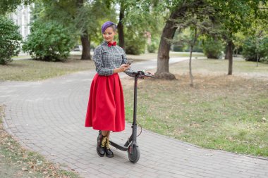 Mor saçlı genç bir kadın parkta elektrikli scooter kullanıyor. Damalı gömlek, uzun kırmızı etek ve papyon lu traşlı bir tapınakla şık kız akıllı telefona mesaj yazıyor. Hipster