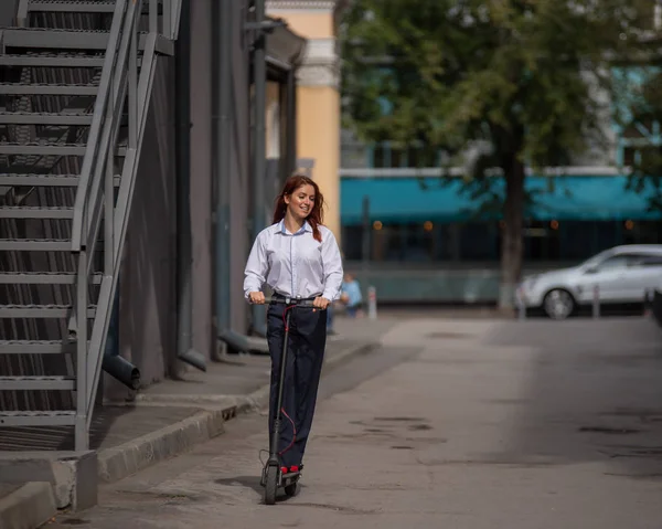 Beyaz gömlekli kızıl saçlı bir kız duvar boyunca elektrikli scooter kullanıyor. Pantolon kostümlü ve kırmızı topuklu ayakkabıgiyen bir iş kadını modern bir arabayla şehirde dolaşıyor. ofiste kıyafet kodu. — Stok fotoğraf