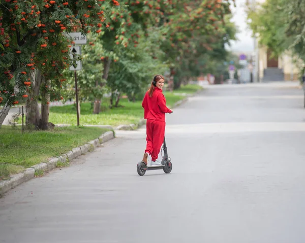 Červenovlasá dívka v červeném obleku řídí elektrický skútr. Mladá žena v příliš velkých šatech jezdí po městě a naslouchá hudbě pomocí bezdrátových sluchátek. Pohled zezadu. — Stock fotografie