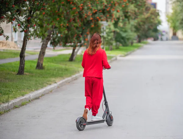 Рыжая девушка в красном спортивном костюме водит электрический скутер. Молодая женщина в огромной одежде разъезжает по городу и слушает музыку с помощью беспроводных наушников. Вид сзади . — стоковое фото