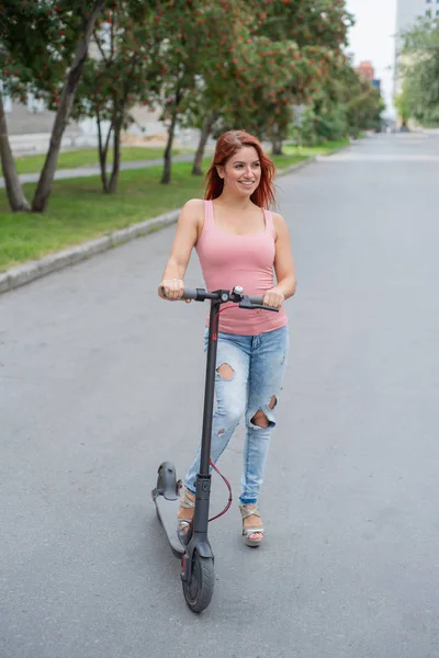 A vörös hajú, fiatal nő a szakadt farmer és a magas sarkú szandálok lovagol egy elektromos robogó az úton. Kényelmes elektromos járművek gyors mozgását a város körül. — Stock Fotó