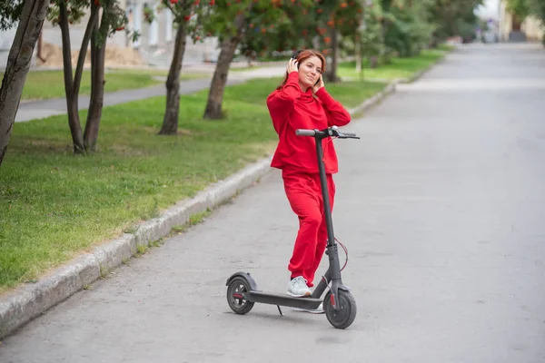 Roodharige meisje in een rood trainingspak drijft een elektrische scooter. Een jonge vrouw in oversized kleding rijdt door de stad op modern transport en luistert naar muziek met behulp van draadloze hoofdtelefoons. — Stockfoto