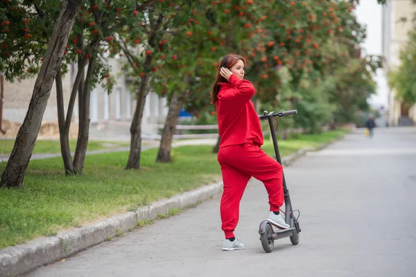 Kırmızı eşofmanlı kızıl saçlı kız elektrikli scooter kullanıyor. Büyük boy giysiler içinde genç bir kadın modern ulaşım şehir etrafında sürmek ve kablosuz kulaklık kullanarak müzik dinler. — Stok fotoğraf