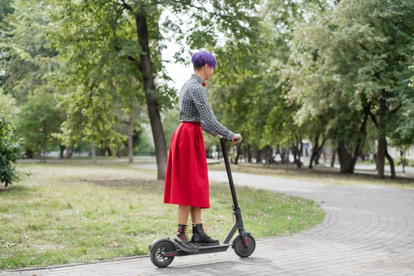 Egy fiatal nő, lila hajú lovagol egy elektromos robogó egy parkban. Egy stílusos lány egy borotvált templom egy kockás ing, egy hosszú vörös szoknya és egy csokornyakkendő a lovaglás a város körül egy modern eszköz. — Stock Fotó