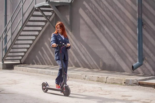 Ceket ve pantolon giyen kızıl saçlı bir kız duvar boyunca elektrikli scooter kullanıyor. Bir pantsuit ve kırmızı yüksek topuklu bir iş kadını modern bir araba ile şehir etrafında sürmek. Ofis çalışanı. Kıyafet kodu — Stok fotoğraf
