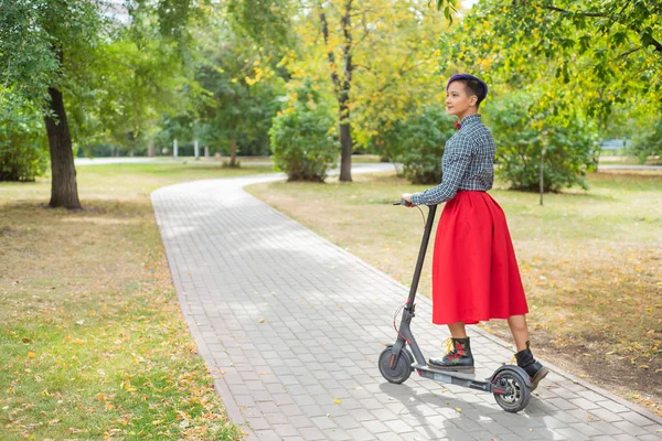 Eine junge Frau mit lila Haaren fährt mit einem Elektroroller in einem Park. ein stylisches Mädchen mit rasiertem Bügel im karierten Hemd, langem roten Rock und Fliege fährt auf einem modernen Gerät durch die Stadt. — Stockfoto