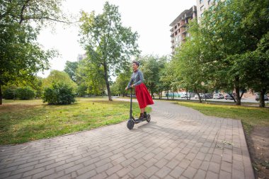 Mor saçlı genç bir kadın parkta elektrikli scooter kullanıyor. Ekose gömlek, uzun kırmızı etek ve papyon bir tıraş tapınak ile şık bir kız modern bir cihaz üzerinde şehir etrafında sürme. 