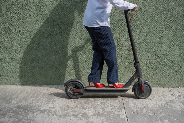 Mujer joven en desgaste formal en tacones rojos de altura está de pie en scooter eléctrico. Primer plano de las piernas femeninas. Una mujer de negocios con un traje de pantalón y zapatos rojos se mueve por la ciudad en un scooter eléctrico . — Foto de Stock