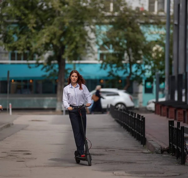 Una ragazza dai capelli rossi con una camicia bianca guida uno scooter elettrico lungo il muro. Una donna d'affari in giacca e cravatta e tacchi rossi gira per la città in un'auto moderna. dress code in ufficio . — Foto Stock