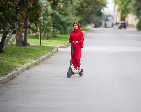 红头发的女孩在红色运动服驾驶电动滑板车。一位身着超大衣服的年轻女子乘坐现代交通工具在城市中四处游荡，使用无线耳机听音乐. — 图库照片