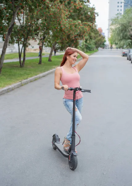 A vörös hajú, fiatal nő a szakadt farmer és a magas sarkú szandálok lovagol egy elektromos robogó az úton. Kényelmes elektromos járművek gyors mozgását a város körül. — Stock Fotó