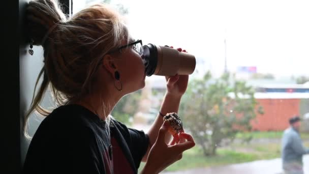 Ein ungewöhnliches Mädchen mit Dreadlocks auf dem Kopf und Tunneln in den Ohren trinkt Kaffee und isst in einem Café einen Donut. eine junge Frau mit Brille frühstückt, während sie bei Regen am Fenster sitzt. — Stockvideo