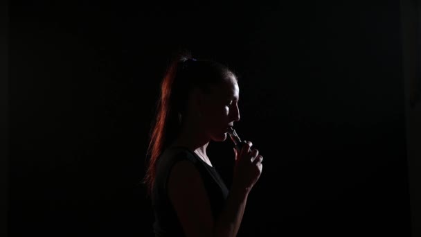 La bruna fuma una sigaretta elettronica su uno sfondo nero, rilascia fumo denso dalla sua bocca. Ritratto di donna che fuma un vaporizzatore, in bilico . — Video Stock