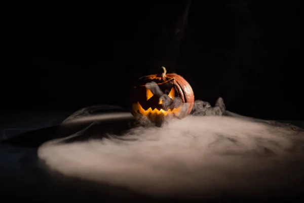 Halloween, apelsin pumpa med en skrämmande lysande ansikte på en mörk bakgrund. Tjock grå rök kommer ut och sprider sig över det svarta bordet. En närbild av en ficklampa på tröskeln till alla helgonen — Stockfoto
