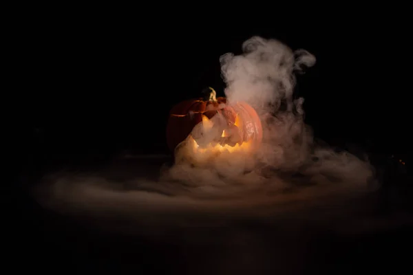 Halloween, calabaza naranja con una cara luminosa de miedo sobre un fondo oscuro. Espeso humo gris sale y se extiende a través de la mesa negra. Un primer plano de una linterna. Soporte de vela decorado . — Foto de Stock