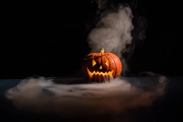 Halloween, citrouille orange avec un visage lumineux effrayant sur un fond sombre. Une épaisse fumée grise se dégage et se propage sur la table noire. Gros plan d'une lampe de poche à la veille de tous les saints — Photo