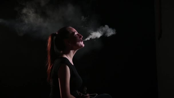 De brunette rookt een elektronische sigaret op een zwarte achtergrond, laat dikke rook uit haar mond ontsnappen. Portret van een vrouw die een vape rookt, zwevend. — Stockvideo