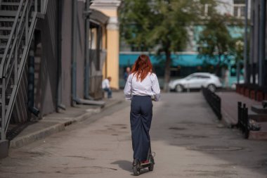 Beyaz gömlekli kızıl saçlı bir kız duvar boyunca elektrikli scooter kullanıyor. Pantolon kostümlü ve kırmızı topuklu ayakkabıgiyen bir iş kadını modern bir arabayla şehirde dolaşıyor. ofiste kıyafet kodu.