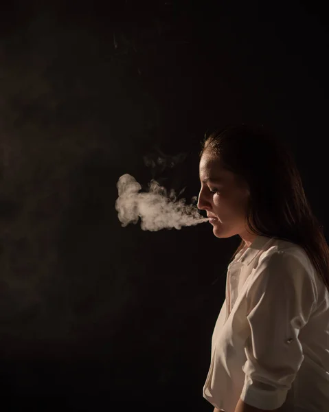 Pensiv brunett i en vit skjorta röker en hookah i mörkret. Porträtt av en kvinna som röker en ånga, svävande. Svart bakgrund. Elektronisk cigarett. Dålig vana. — Stockfoto