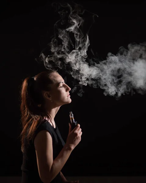 Брюнетка курить електронну сигарету на чорному тлі, вивільняючи густий дим з рота. Портрет жінки, яка курить вейп, що нависає . — стокове фото