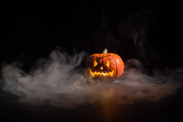 Halloween, orange pumpkin with a scary luminous face on a dark background. Thick gray smoke comes out and spreads across the black table. A close-up of a flashlight on the eve of all the saints — Stock Photo, Image