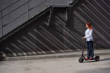Beyaz gömlekli kızıl saçlı bir kız duvar boyunca elektrikli scooter kullanıyor. Pantolon kostümlü ve kırmızı topuklu ayakkabıgiyen bir iş kadını modern bir arabayla şehirde dolaşıyor. Scooter'da iş kadını.
