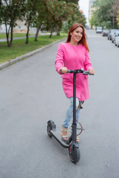 Rudowłosy młoda kobieta w zgranej dżinsy i sandały na obcasie jeździ elektryczny skuter na drodze. Dziewczyna w różowej marynarce szybko porusza się po mieście na wygodnym transporcie elektrycznym. — Zdjęcie stockowe