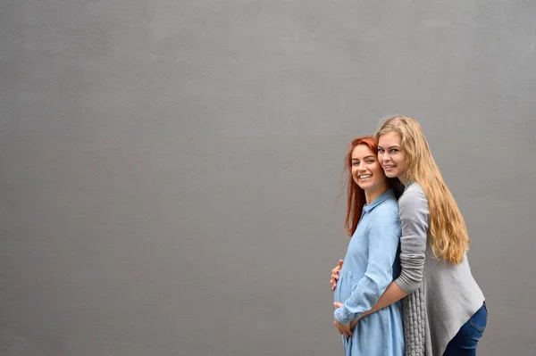 Relaties met hetzelfde geslacht. Een roodharige zwangere vrouw in een denim jurk staat tegen een grijze muur, haar hand rust onder haar buik. Haar vrouw zachtjes omhelsde de aanstaande moeder van achteren. — Stockfoto