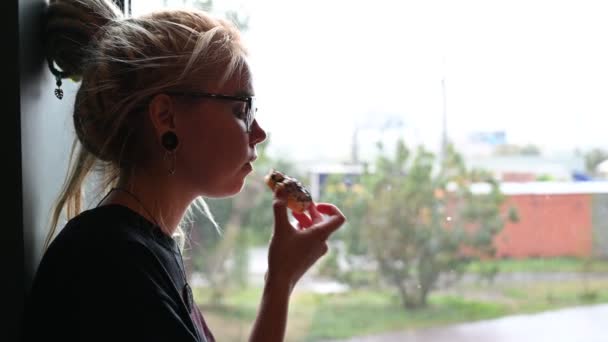 Una chica inusual con rastas en la cabeza y túneles en los oídos bebe café y se come un donut en un café. Una joven con gafas está desayunando sentada junto a la ventana bajo la lluvia. . — Vídeo de stock