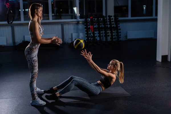 The blonde from the floor throws a stuffed ball to the coach. Circular functional training with a trainer. A woman working on her abs with a weighted ball