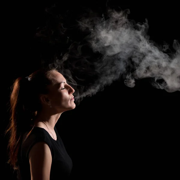 De brunette rookt een elektronische sigaret op een zwarte achtergrond, laat dikke rook uit haar mond ontsnappen. Portret van een vrouw die een vape rookt, zwevend. — Stockfoto