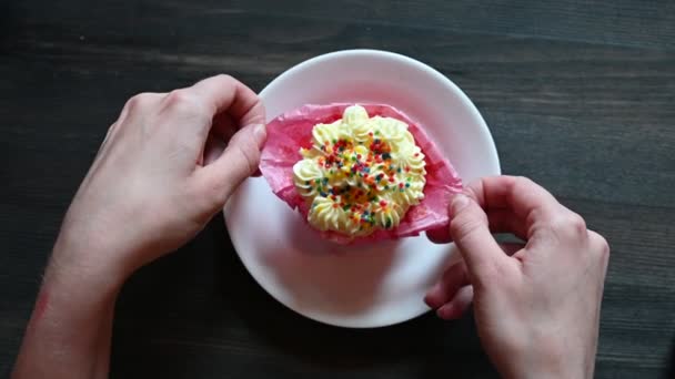 女性はカフェでクリームとコンフィティで甘いカップケーキを食べています。大きな女性の手はカップケーキを展開.ケーキ付きの白いプレートのトップビュー. — ストック動画