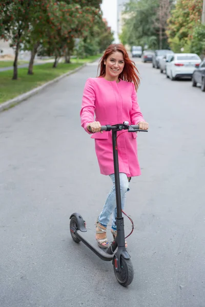 Μια κοκκινομάλλα νεαρή γυναίκα με σκισμένα τζιν και τακούνια σανδάλια βόλτες με ένα ηλεκτρικό σκούτερ στο δρόμο. Ένα κορίτσι σε ένα ροζ μπουφάν κινείται γρήγορα γύρω από την πόλη σε μια βολική ηλεκτρική μεταφορά. — Φωτογραφία Αρχείου